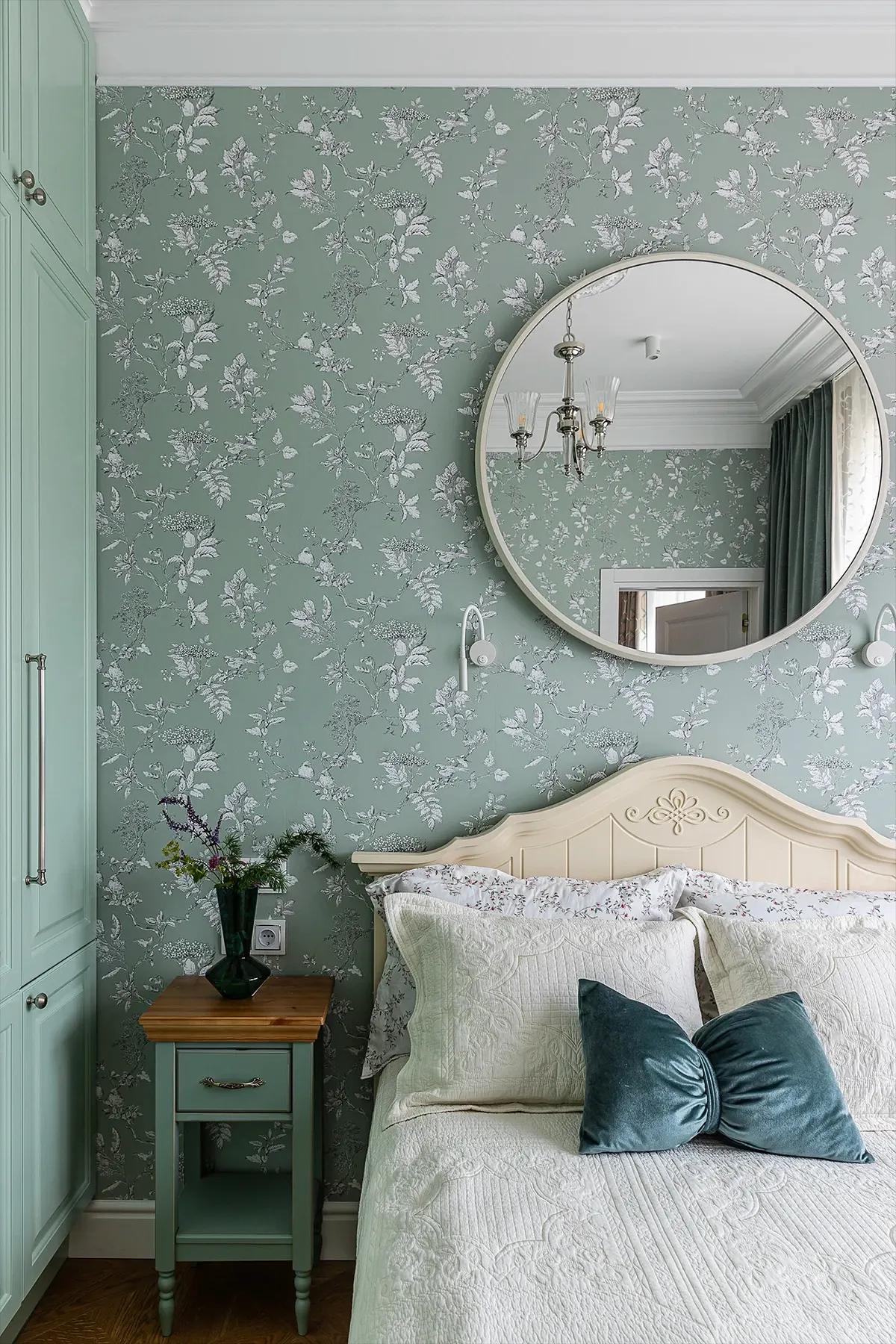 Уютная светлая спальня в зеленых тонах с круглым зеркалом на стене и с цветами в вазе на прикроватной тумбочке