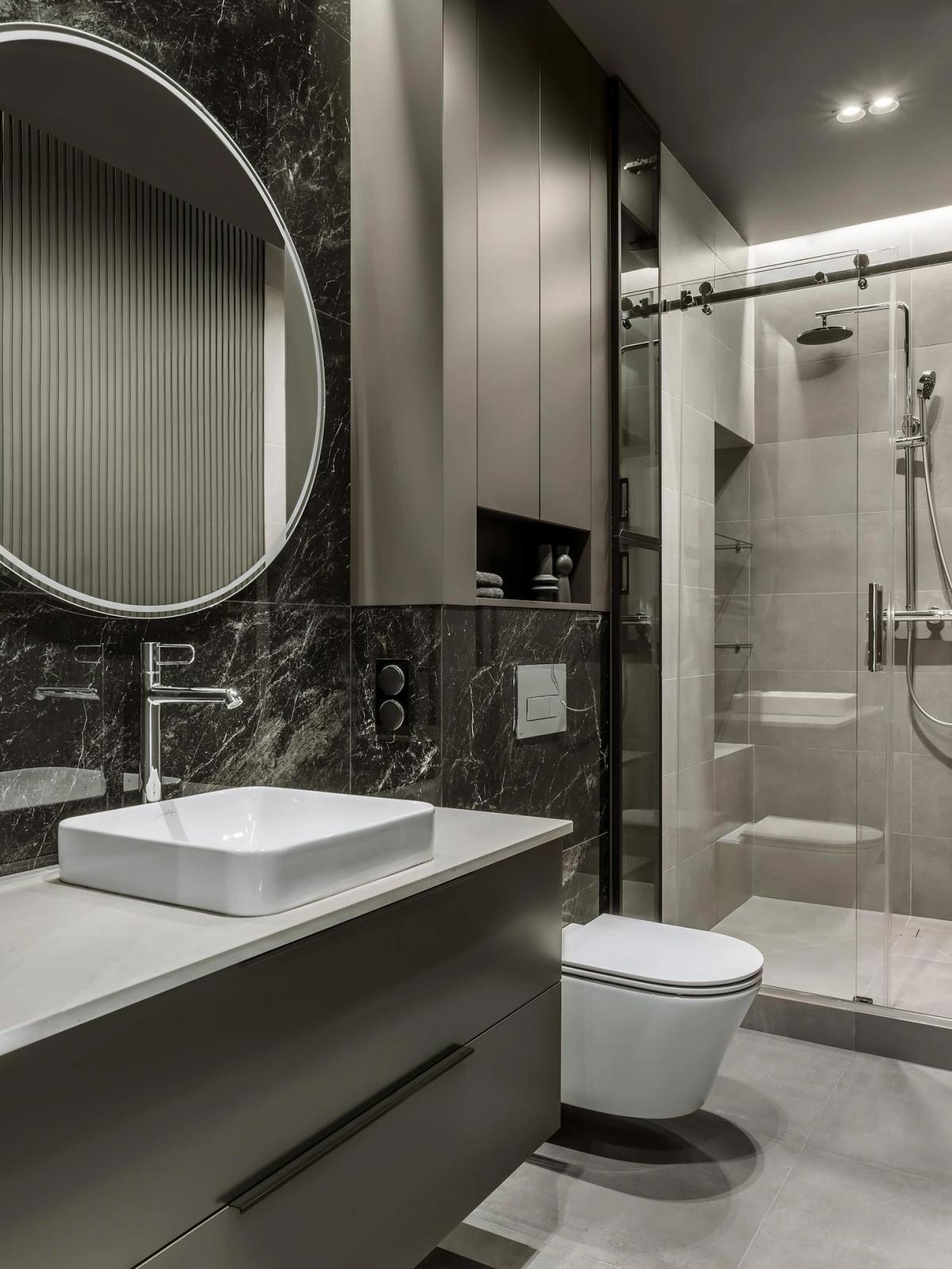 Минималистичная ванная комната с мраморными стенами, круглым зеркалом, белой раковиной, душем за стеклом