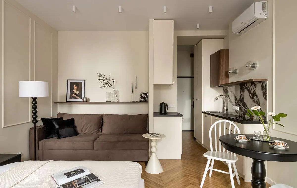 Интерьер современной квартиры-студии с коричневым диваном, небольшим обеденным столом со стульями, мини-кухней с мраморным фартуком и паркетом в "елочку"