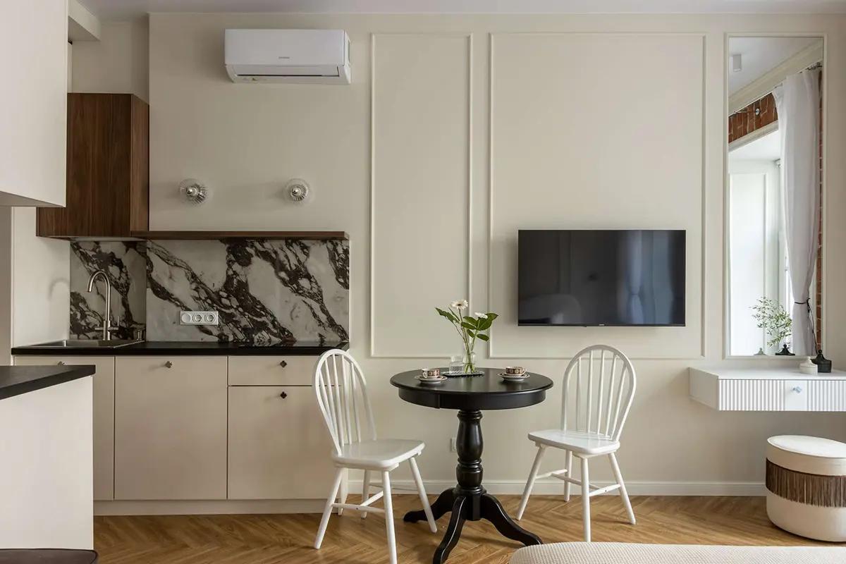 Кухонная зона в студии с мраморным фартуком, черным обеденным столом, белыми стульями, телевизором на стене и цветами в вазе