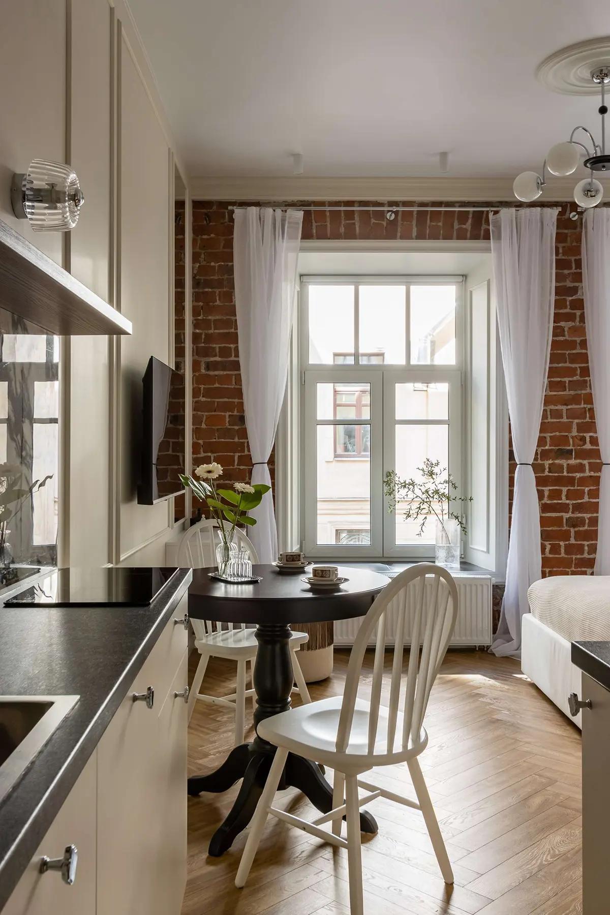 Кухня с высоким окном, кирпичной стеной, тюлью, черным столом и белыми стульями