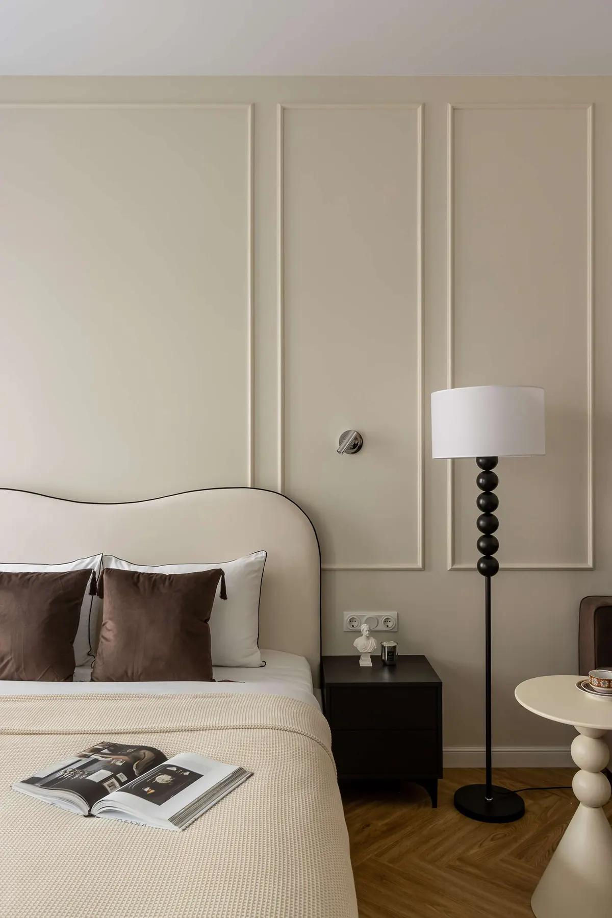 Элегантный уголок спальни в компактной квартире с белой кроватью, коричневыми подушками, черной тумбочкой, торшером и открытым журналом на покрывале