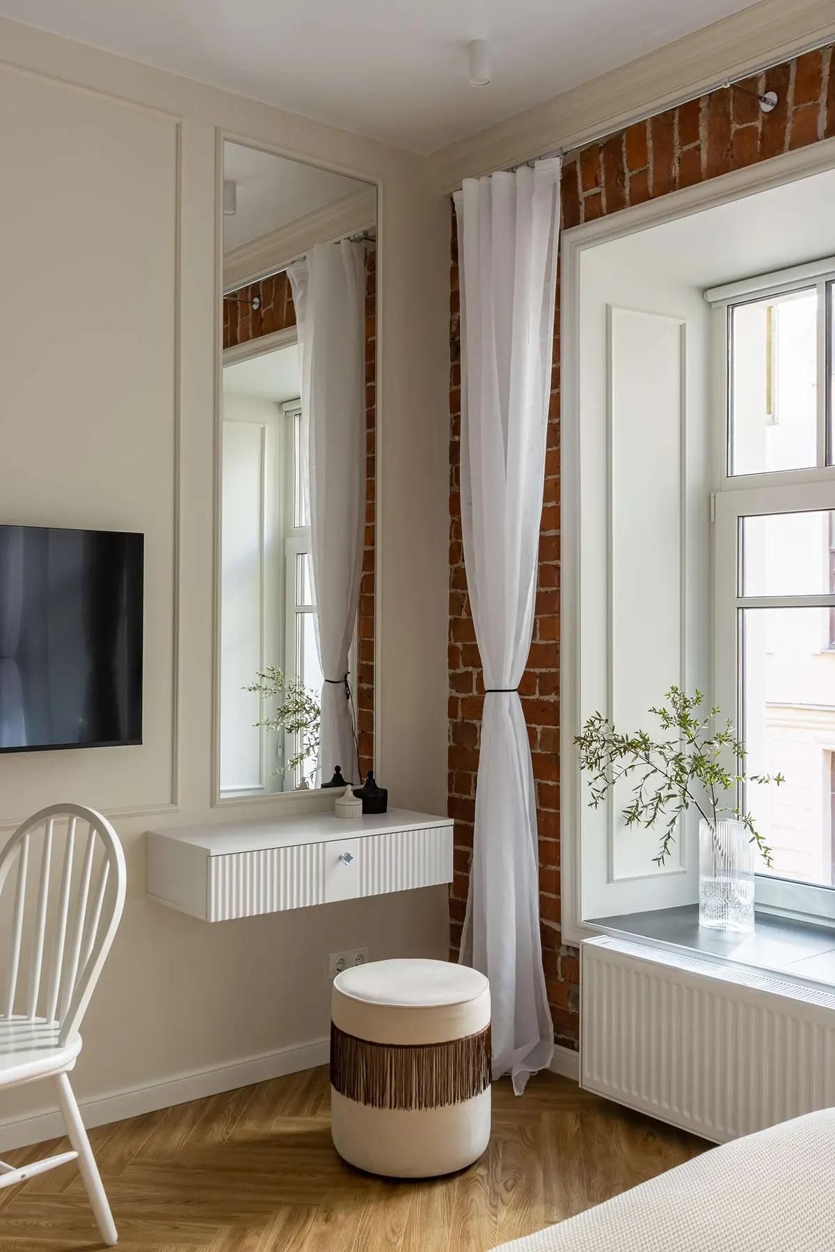 Уголок у окна в квартире с настенным телевизором, белым туалетным столиком, высоким зеркалом, пуфом с бахромой и растениями в вазах на подоконнике