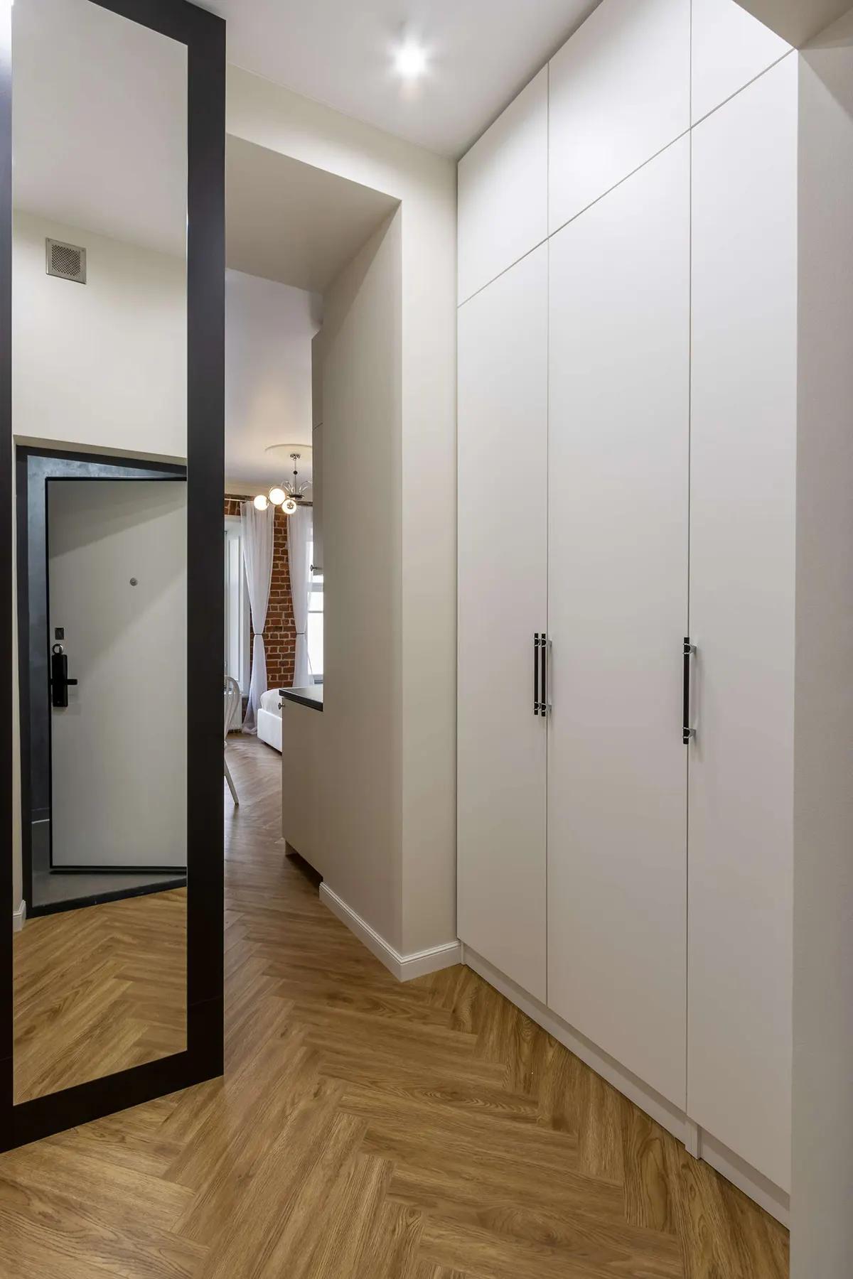 Узкий коридор с белыми шкафами, зеркалом и ламинатом в елочку