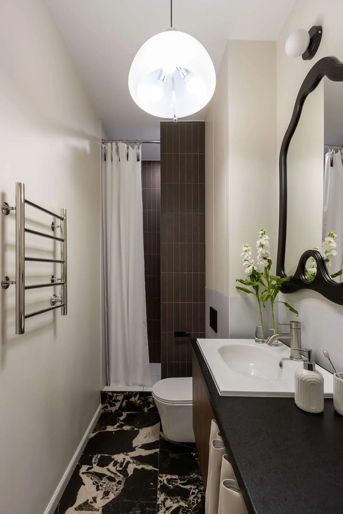 Компактная ванная комната с душем за шторкой, белыми цветами на умывальнике, черным зеркалом, полотенцесушителем и мраморным полом в черно-белых тонах