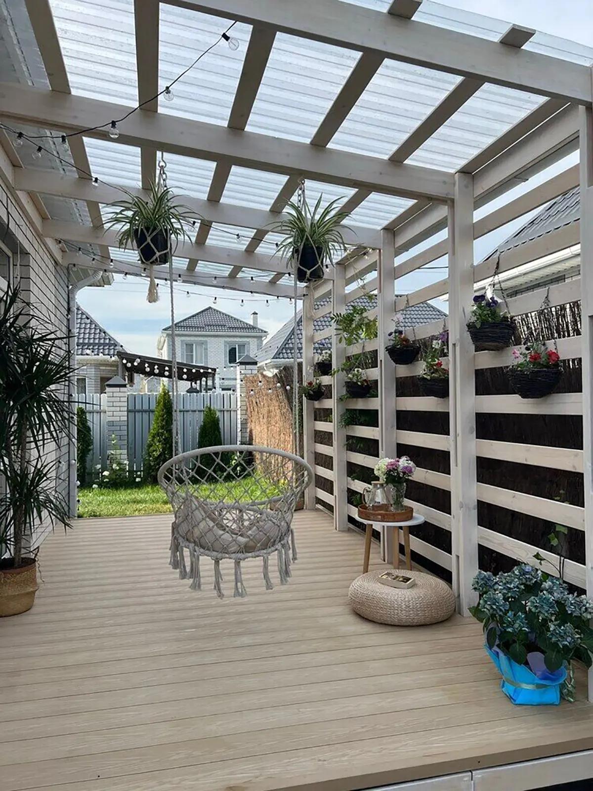 Терраса с деревянным настилом, прозрачной крышей, висящими растениями, гирляндой лампочек и качающимся креслом