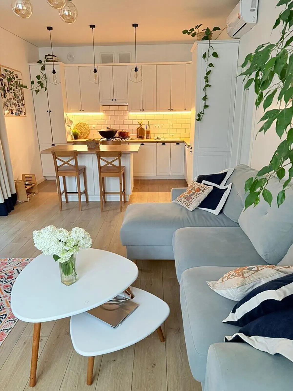 Кухня-гостиная с белыми шкафами, деревянной барной стойкой, серым диваном и белым журнальным столиком