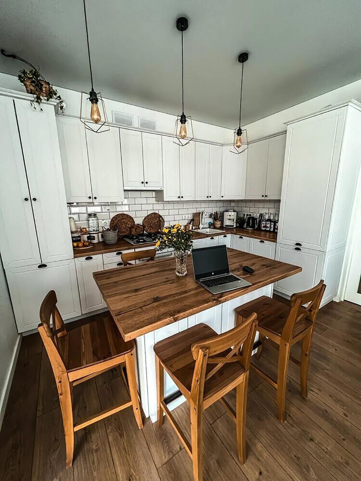 Кухня с белыми шкафами, деревянным кухонным островом, стульями, ноутбуком, висящими лампами