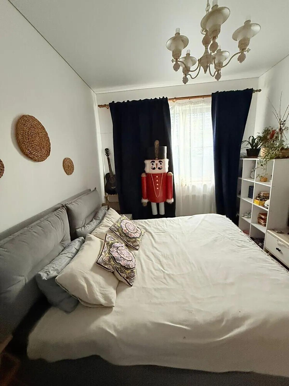 Спальня с белыми стенами и темными шторами. Кровать с серым изголовьем. Щелкунчик в человеческий рост