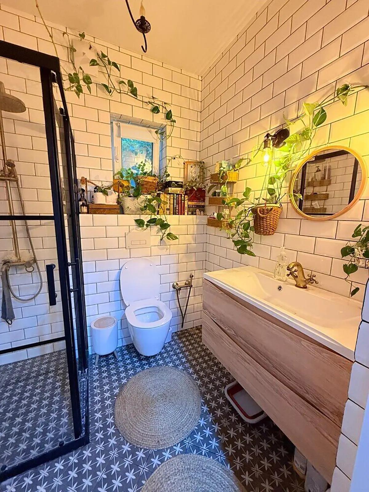 Ванная комната с белой плиткой, висящими растениями, лампочками, круглым зеркалом, раковиной, унитазом и душевой кабиной