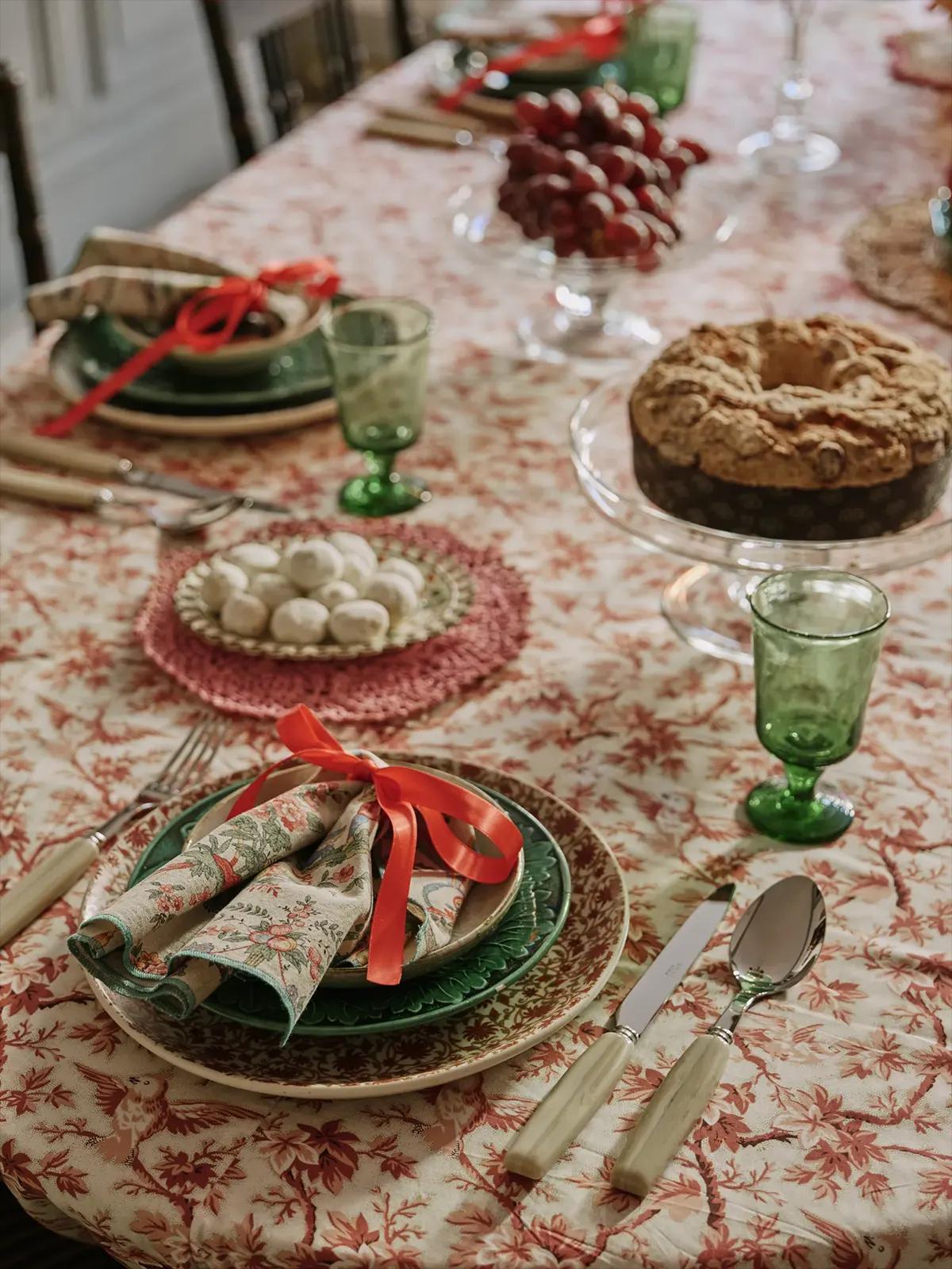 Праздничный стол с тортом, виноградом, белыми шариками, зелёными бокалами, цветочной салфеткой с красной лентой и приборами на скатерти