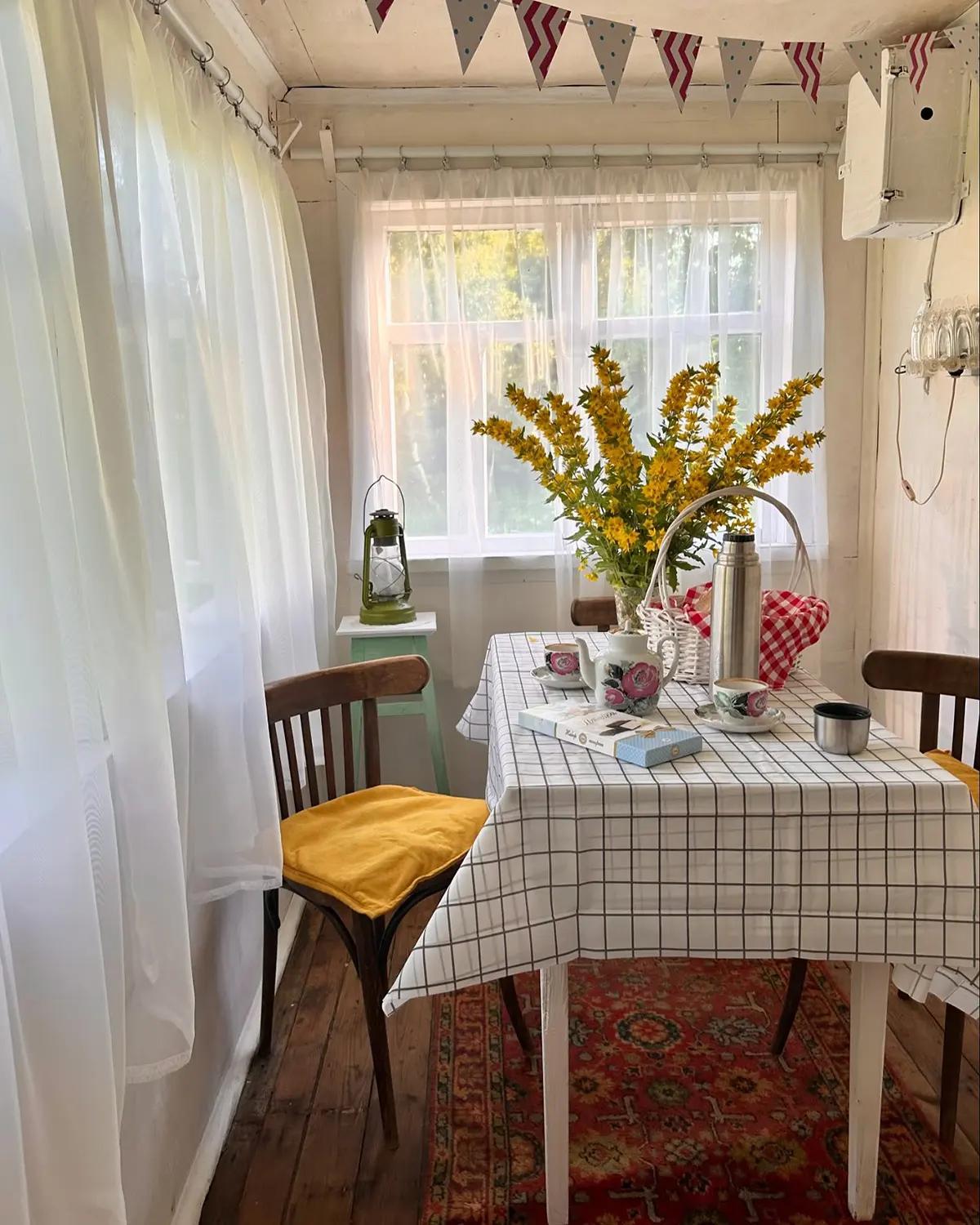 Белый стол с клечатой скатертью, полупрозрачные занавески на окне, гирлянда из треугольных флажков на светлой веранде