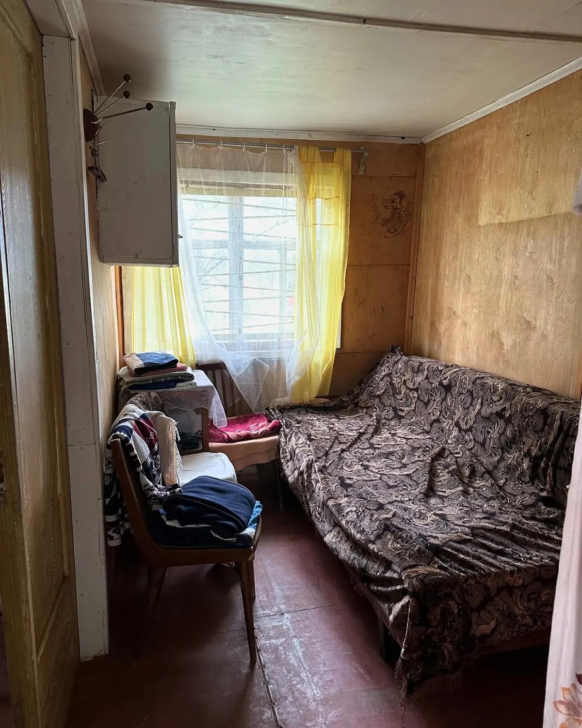 Спальня в доме до ремонта с деревянными полом и стенами и диваном с пледом темной раскраски