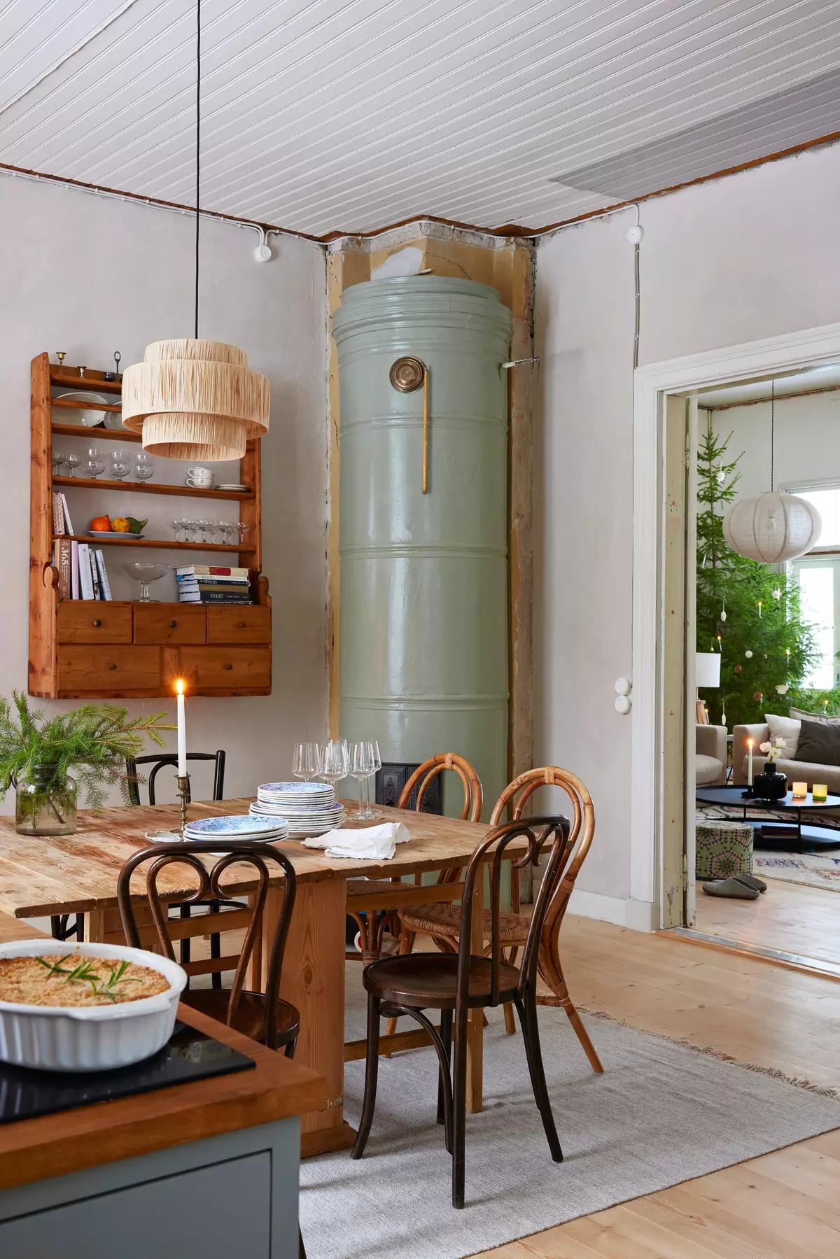 Обеденный деревянный стол, стульями, полкой с посудой, зеленой печью в углу, и видом на елку в гостиной