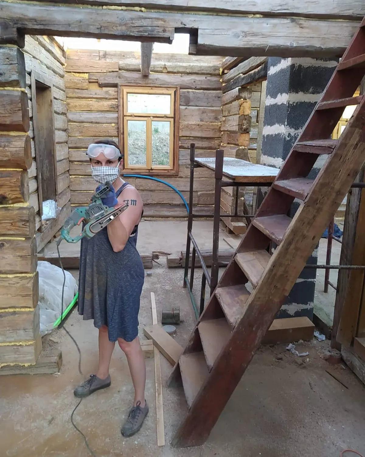 Женщина в защитной маске и очках шлифует стену в деревянном доме