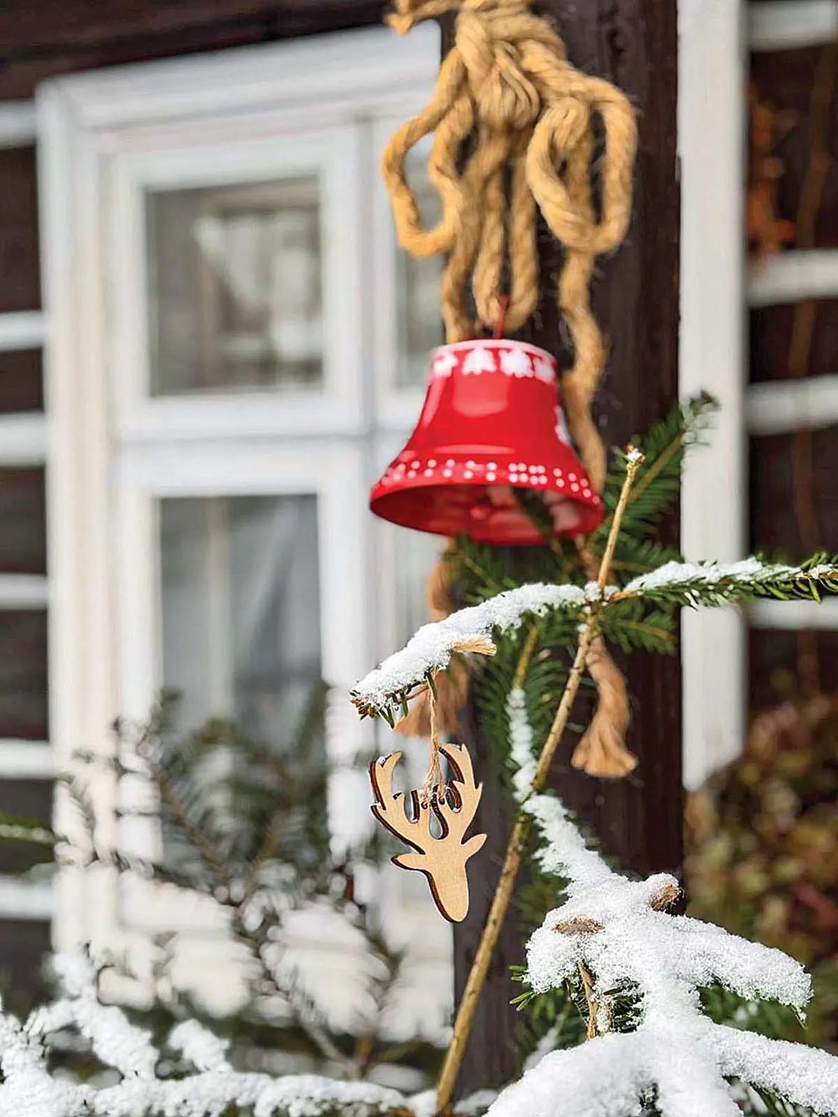 Красный колокольчик и деревянный олень на веревке с заснеженной еловой веткой на фоне окна