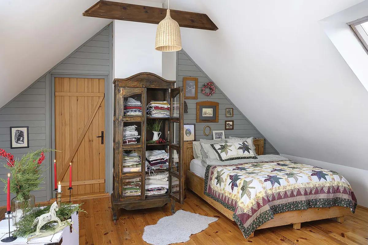Уютная мансардная спальня с наклонными серыми стенами, антикварным деревянным шкафом с бельем, лоскутным одеялом на кровати, свечами и настенными картинами.