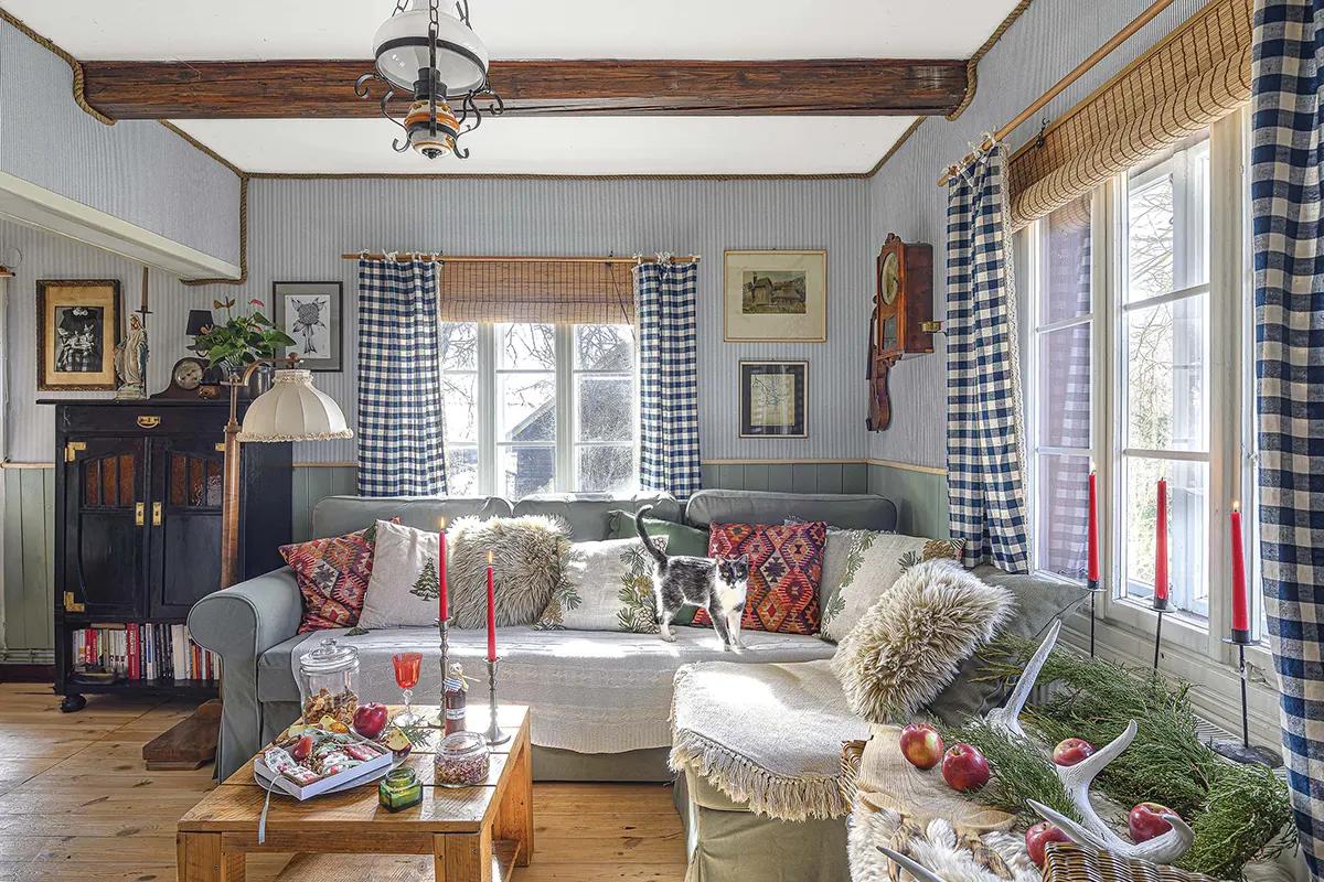 Гостиная в кантри-стиле с серым диваном, яркими подушками, кошкой на диване, журнальным столиком, клетчатыми шторами и деревянным полом