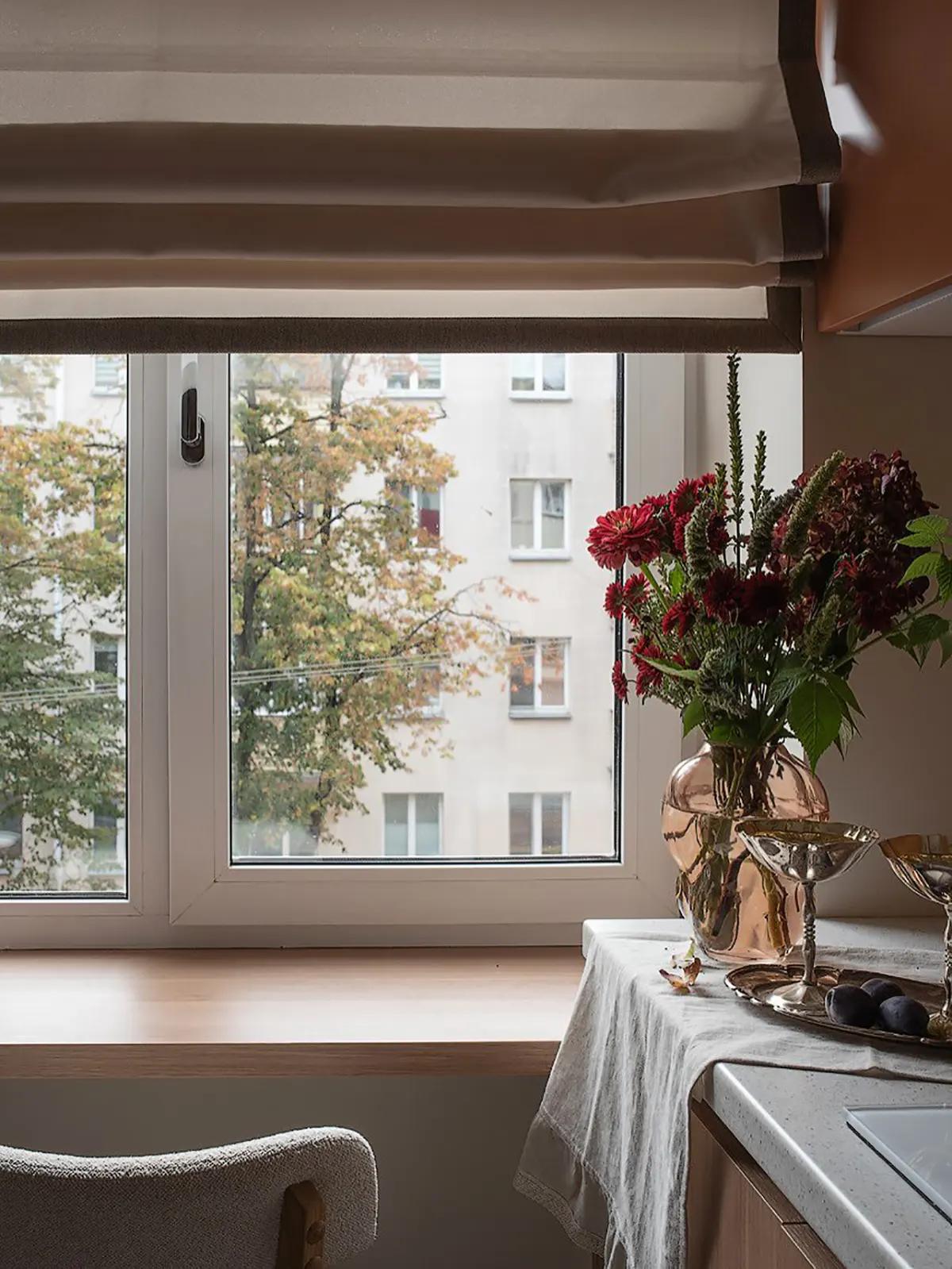 Кухонное окно со столом-подоконником с видом на осенний Минск. Букет красных цветов в бледно-розовой прозрачной вазе на столешнице