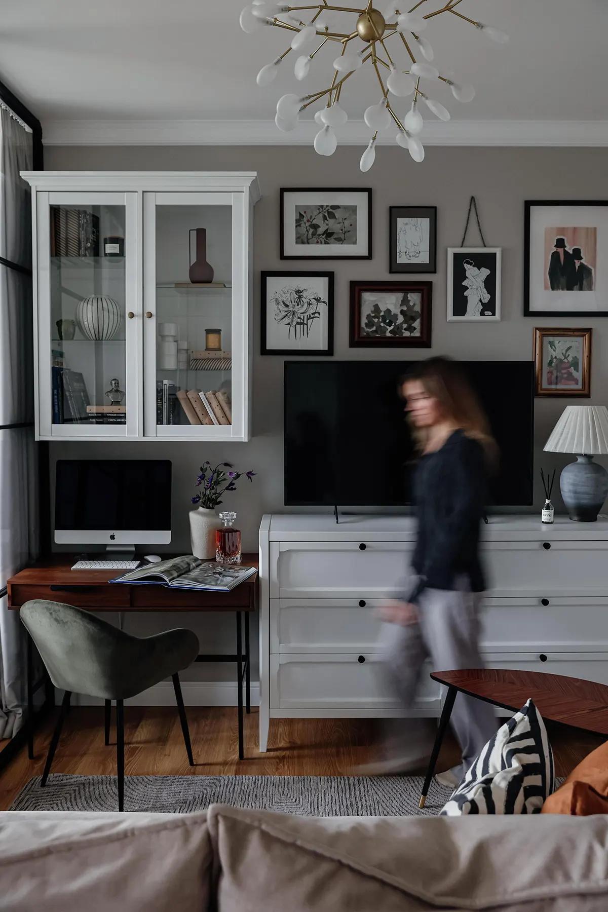 Комната с белым шкафом-витриной, комодом, телевизором, рабочим столом, размытым силуэтом человека и галереей картин на стене