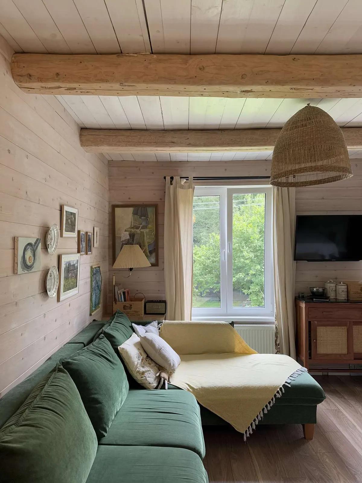 Зеленый угловой диван в деревянной гостиной с картинами на стене, окном, телевизором и пледом