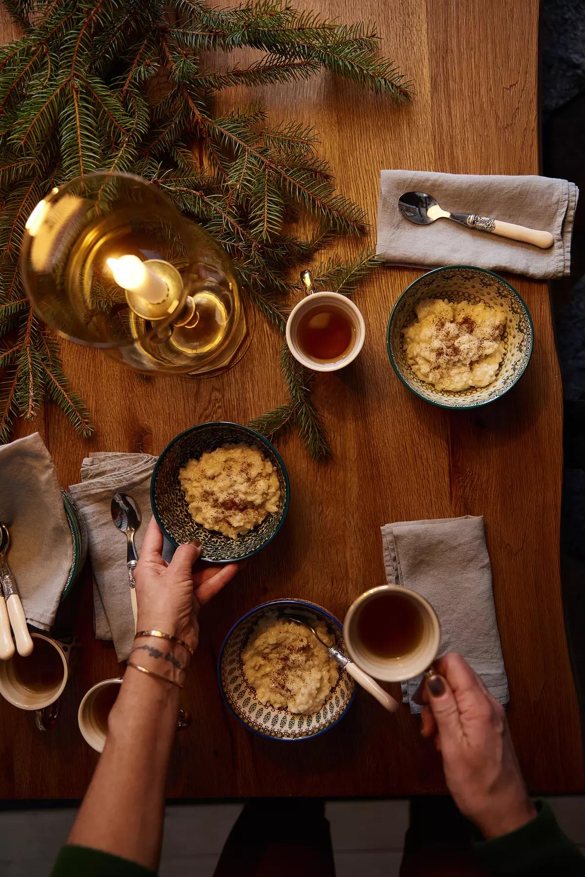Стол с мисками каши, чашками чая, руками, держащими посуду, и еловыми ветками