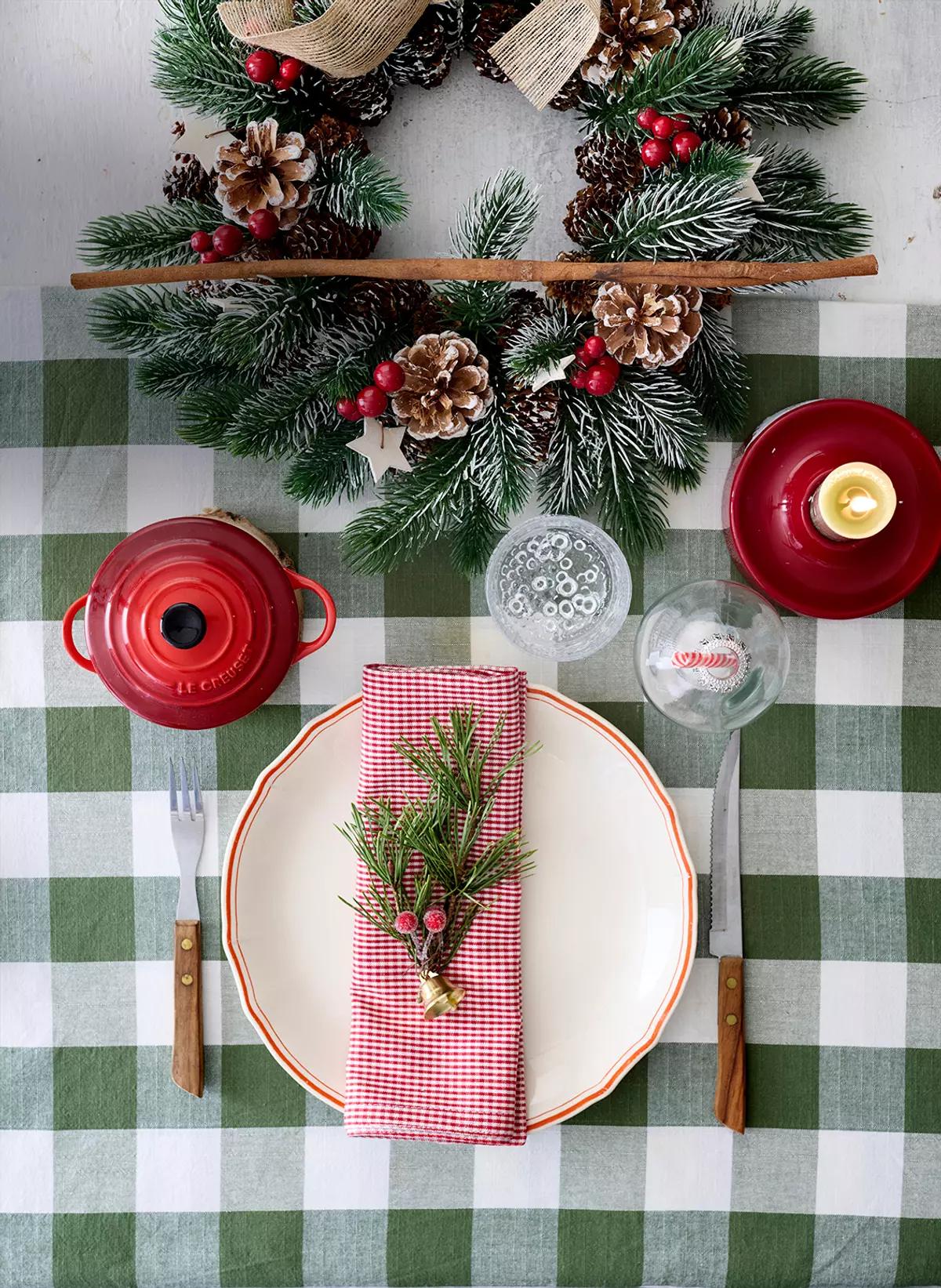 Сервировка на зелёной клетчатой скатерти с венком, белой тарелкой, красной салфеткой и еловой веткой