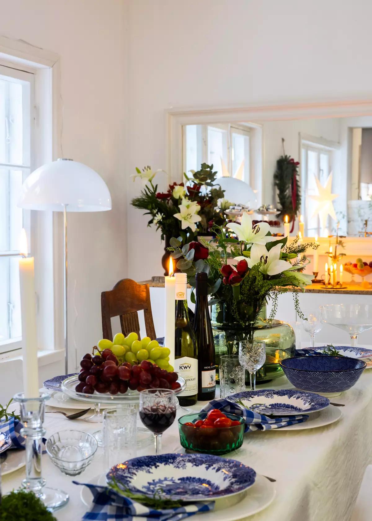 Праздничный стол с белой скатертью, синими тарелками, цветами в вазе, вином, фруктами, 
