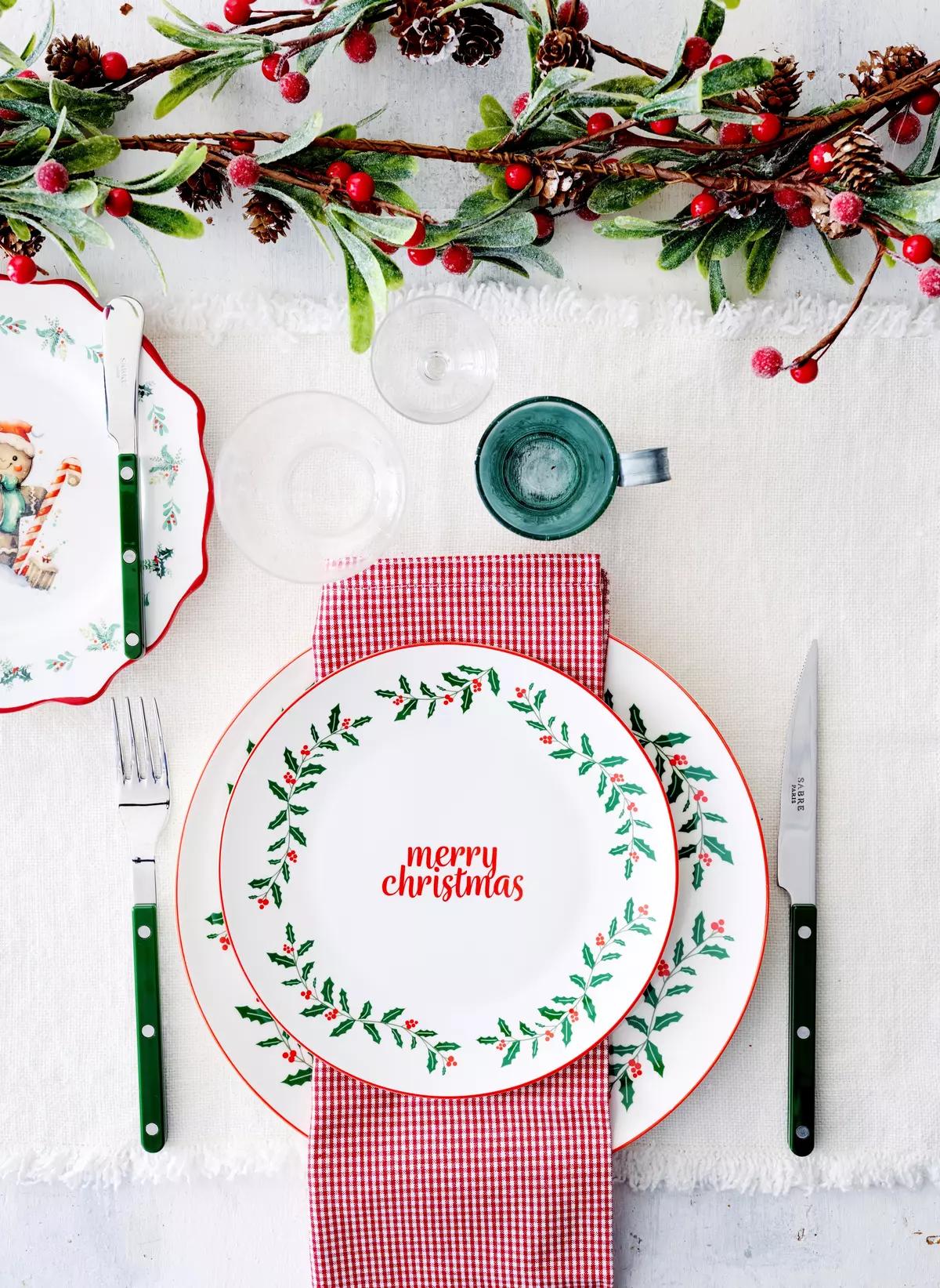 Рождественская сервировка стола с тарелкой "Merry Christmas", красной клетчатой салфеткой, столовыми приборами и веточкой с ягодами