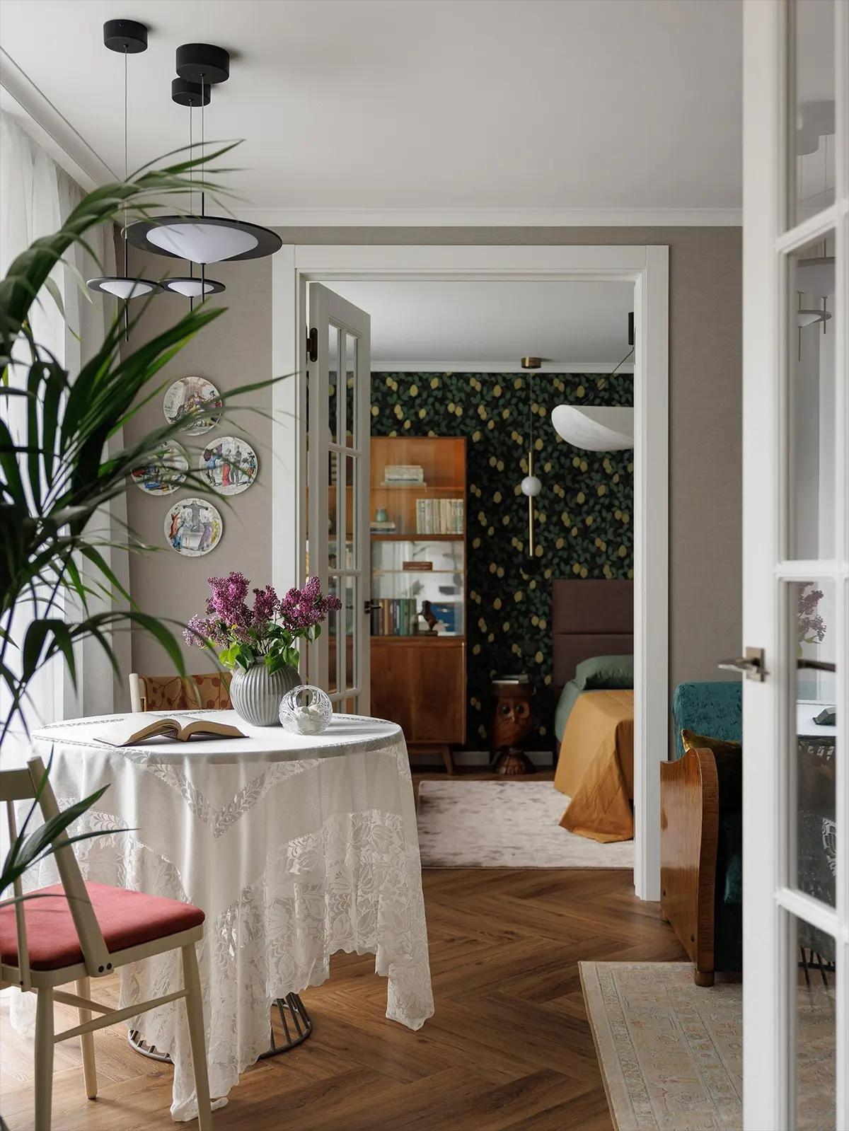 Круглый обеденный стол с белой кружевной скатертью на фоне спальни. На столе ваза с сиренью и открытая книга