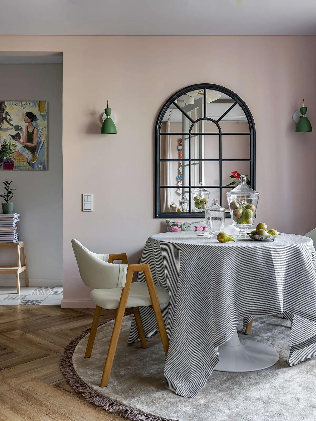 Арочное зеркало в черной раме на бледно-розовой стене в гостиной. Круглый стол, накрытый полосатой скатертью