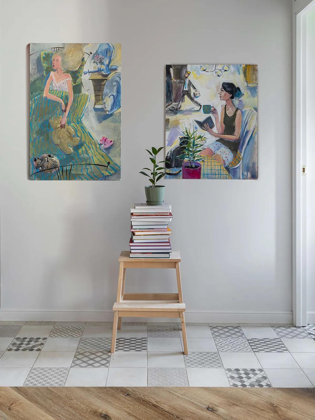 Прихожая в хрущевке с абстрактными картинами на белой стене, стопкой книг на деревянном табурете