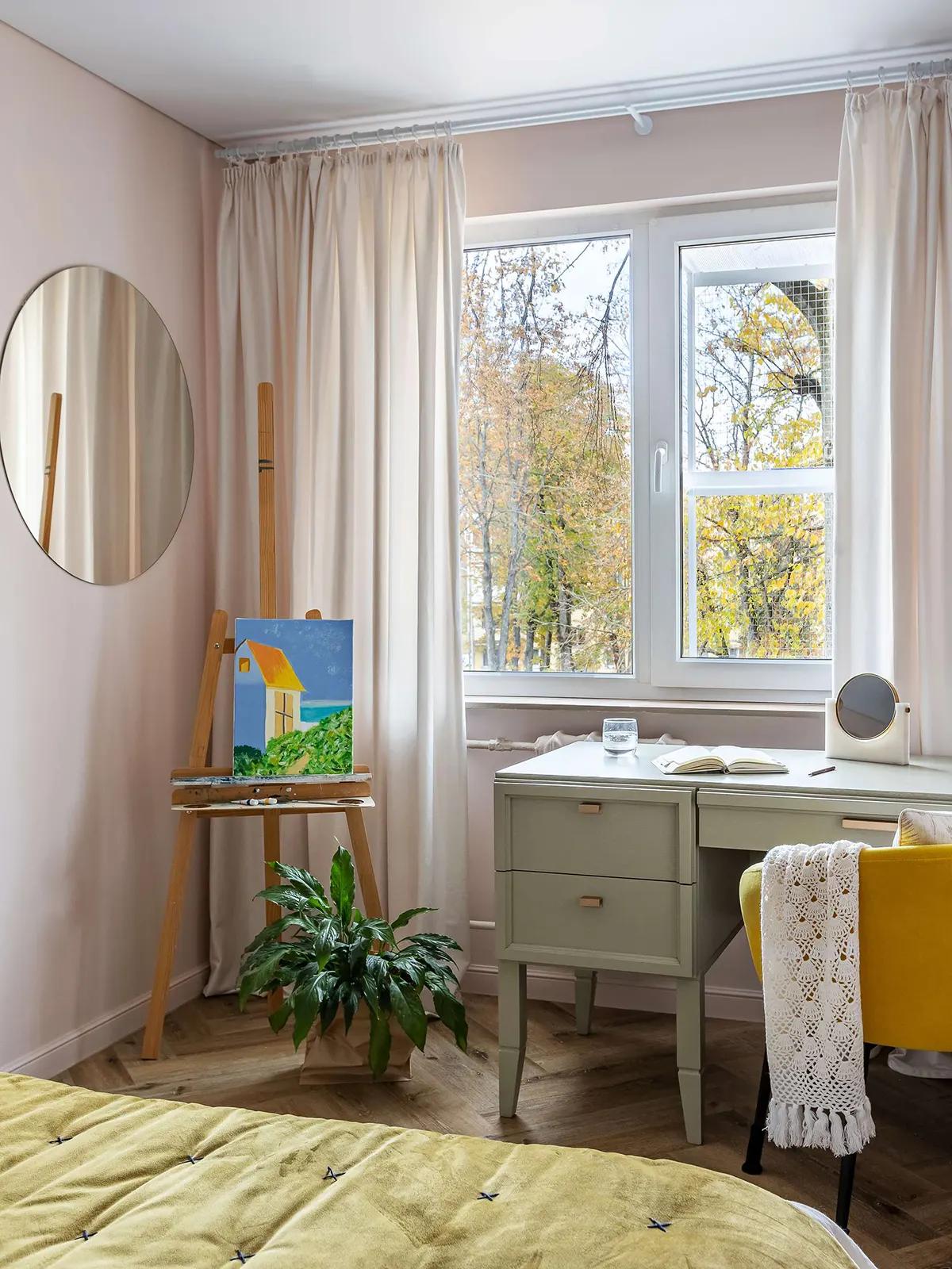 Спальня с мольбертом у окна в хрущевке, стол с блокнотом, желтое кресло с пледом, растение и вид на осенние деревья
