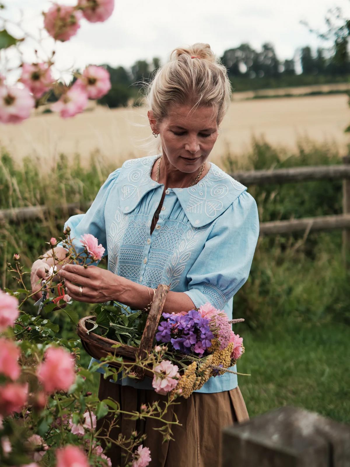 Александра Толстая в голубой блузке собирает цветы в корзину