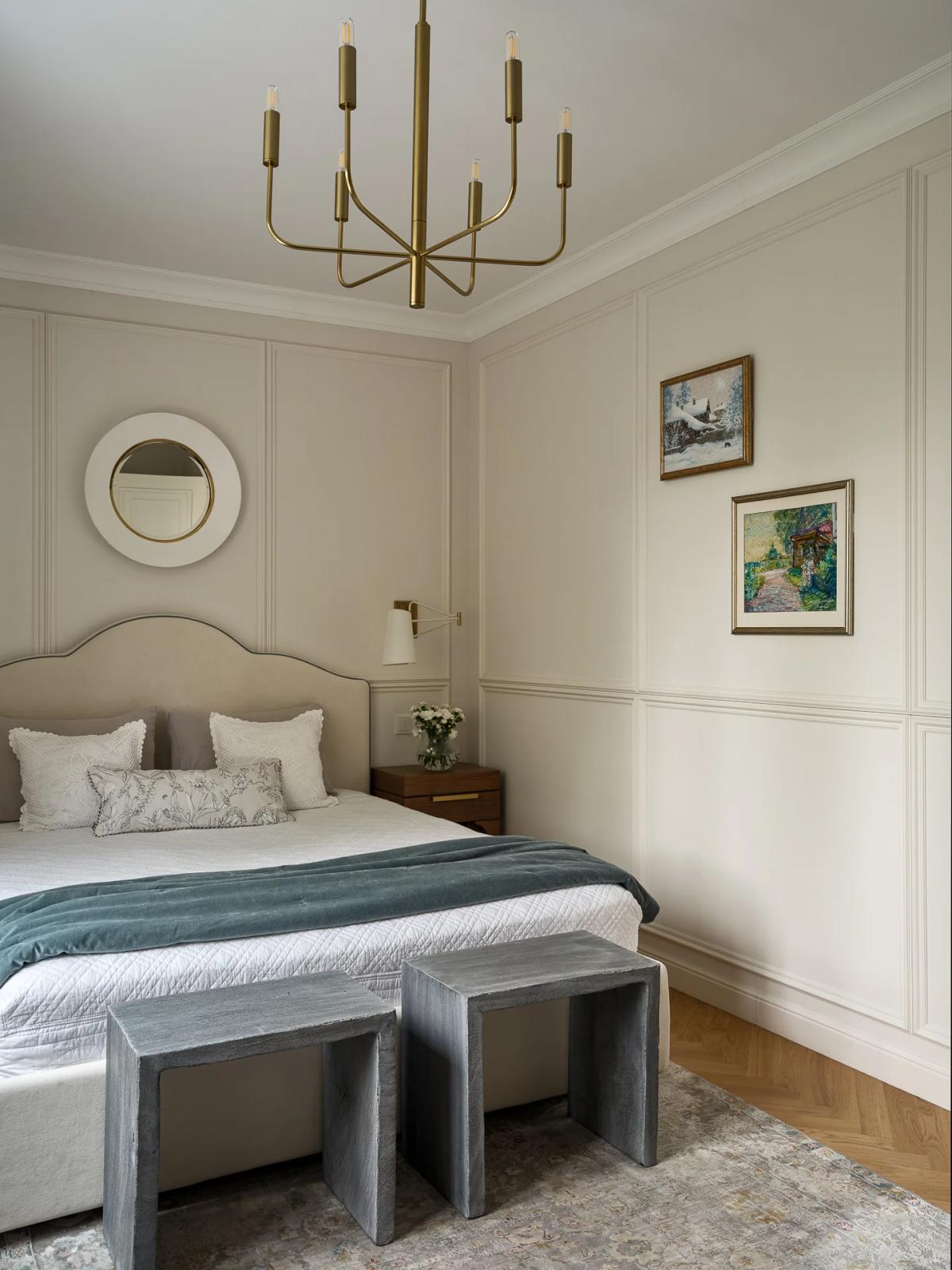 Уютная спальня с бежевым изголовьем кровати, круглым белым зеркалом, золотой настенной лампой, белыми цветами в вазе